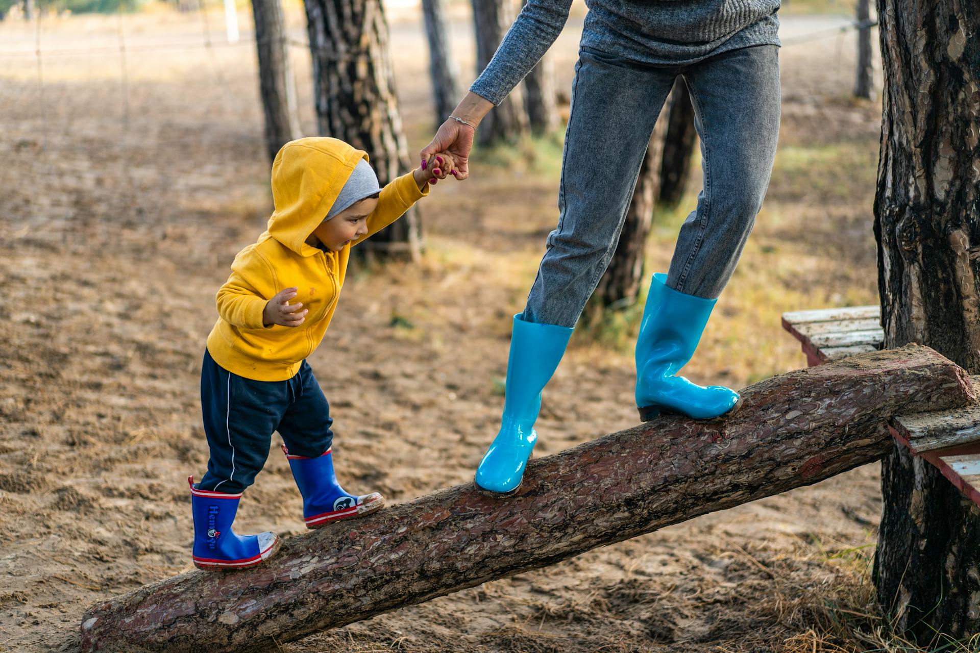 Un enfant avec des bottes en caoutchouc bleu et rouge et un sweat à capuche marche sur un tronc d'arbre couché avec l'aide d'un adulte